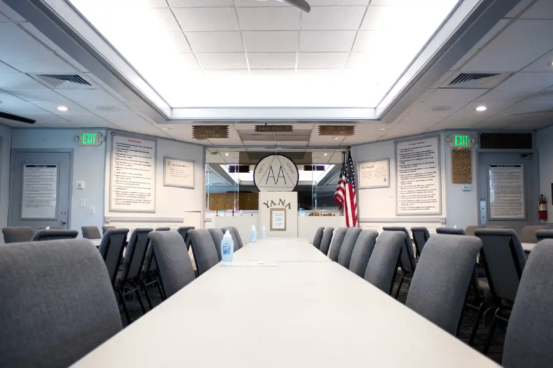 Interior of a meeting room at Y.A.N.A. with long conference tables, gray chairs, framed posters on the walls, AA signage, and an American flag near the podium